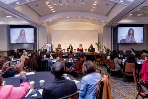 Four women panelists discuss health equity on stage; audience listens in a modern, well-lit conference room.